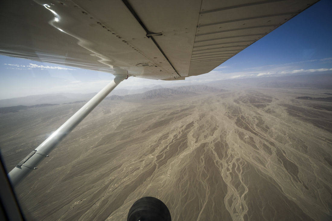 Vliegtuigvleugel van een klein vliegtuigje gezien vanaf binnen met erachter de droge grond rondom Nazca