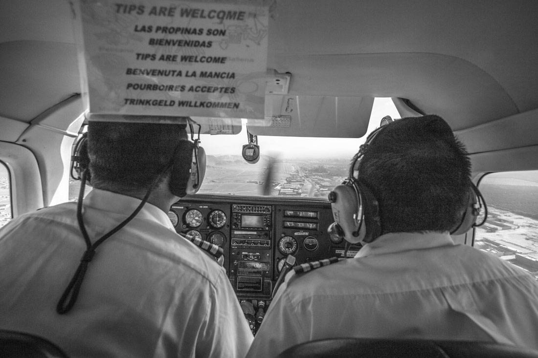 Zwartwitfoto van twee piloten die voorin een klein vliegtuigje zitten met ervoor een papiertje waarop in meerdere talen staan 'tips are welcome'
