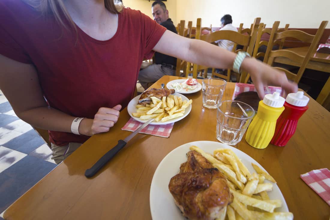 Manouk reikt naar een fles saus en op de tafel staan twee borden met kip en frietjes.