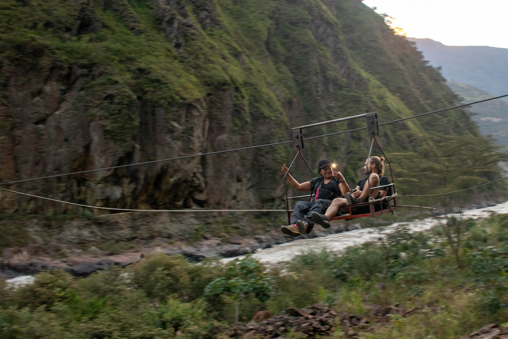 Handkabelbaan tijdens de Inka Jungle Trek: twee mensen zitten op een plateautje, dat een local met de hand naar de overkant trekt.