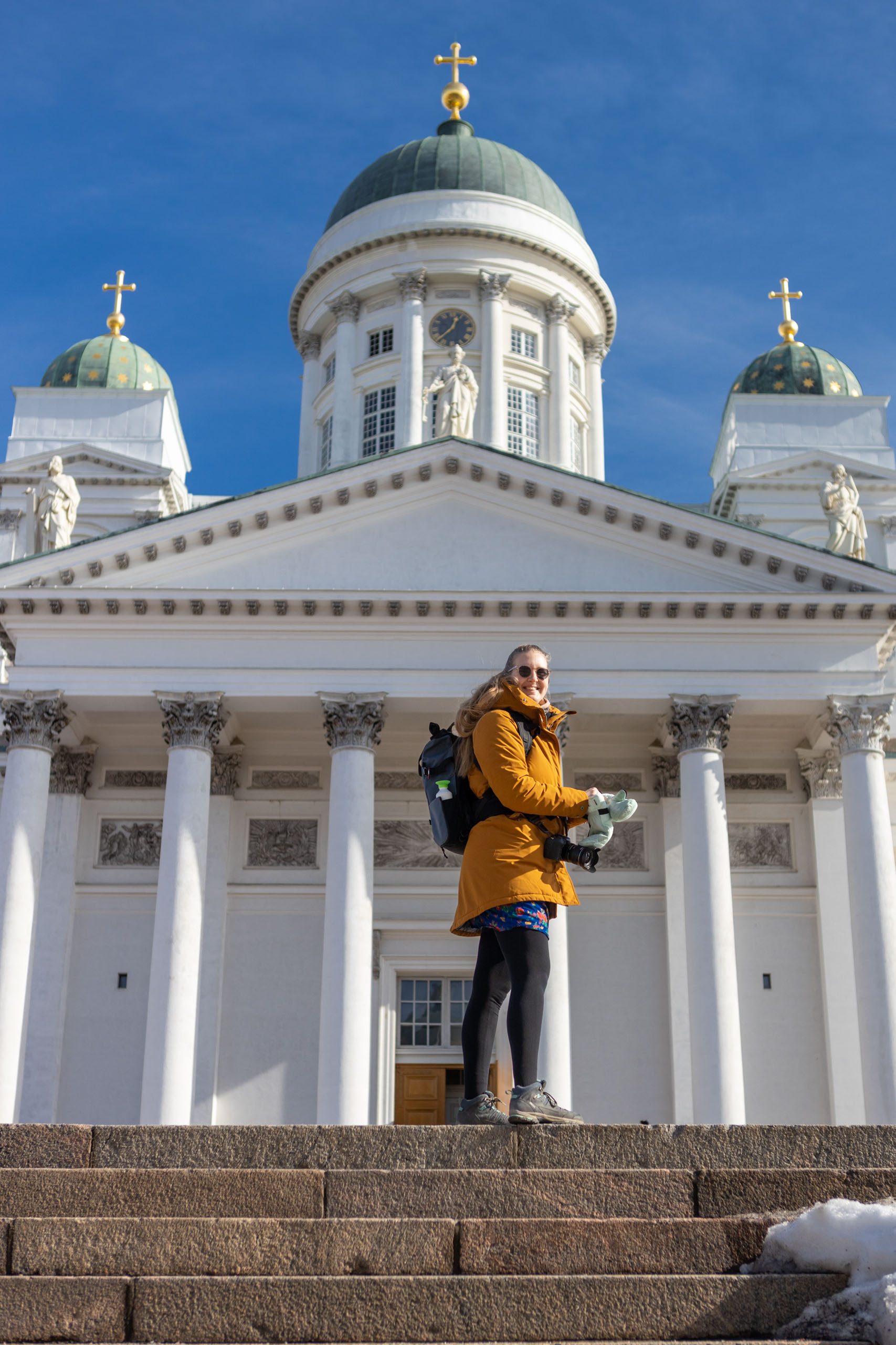 Manouk in gele jas op de trap voor de witte Domkerk Tuomiokirkko in Helsinki met blauwe lucht erachter