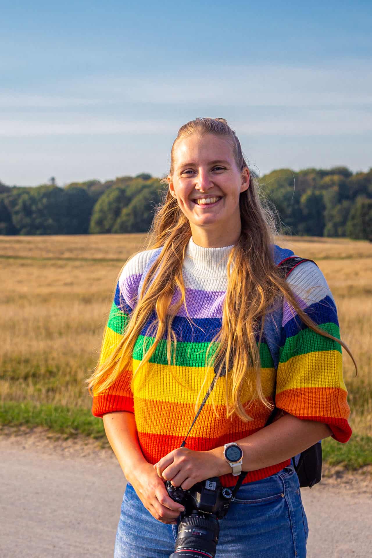 Manouk van Groetjes uit Verweggistan staat in een regenboogtrui voor een geel veld in een park in Denemarken met camera in haar handen