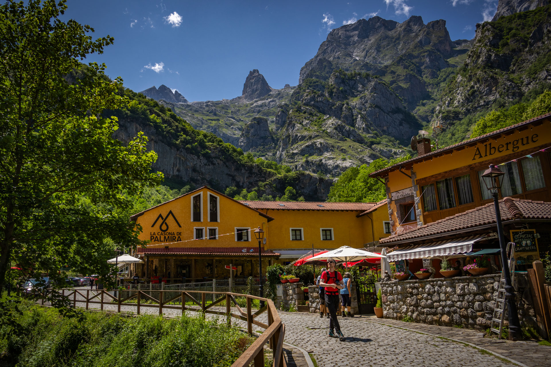 Pauzeren van de wandeling Ruta del Cares in Picos de Europa in Spanje, je ziet een geel restaurant waar Hugo voor loopt over een pad met bergen en groen tegen een blauwe lucht.