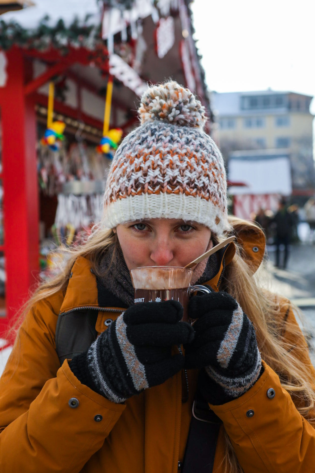 Manouk drinkt een warme chocolademelk uit een glas met een muts op en handschoenen aan met achter haar een kraam op de kerstmarkt in Koblenz