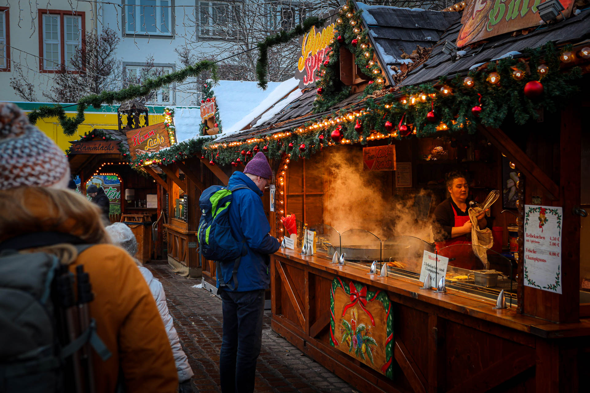 Manouk met gele jas en muts loopt langs kraampjes waar je eten kunt kopen op de kerstmarkt in Koblenz, met lichtjes boven de kraam en rook die uit de kramen komt door de kou