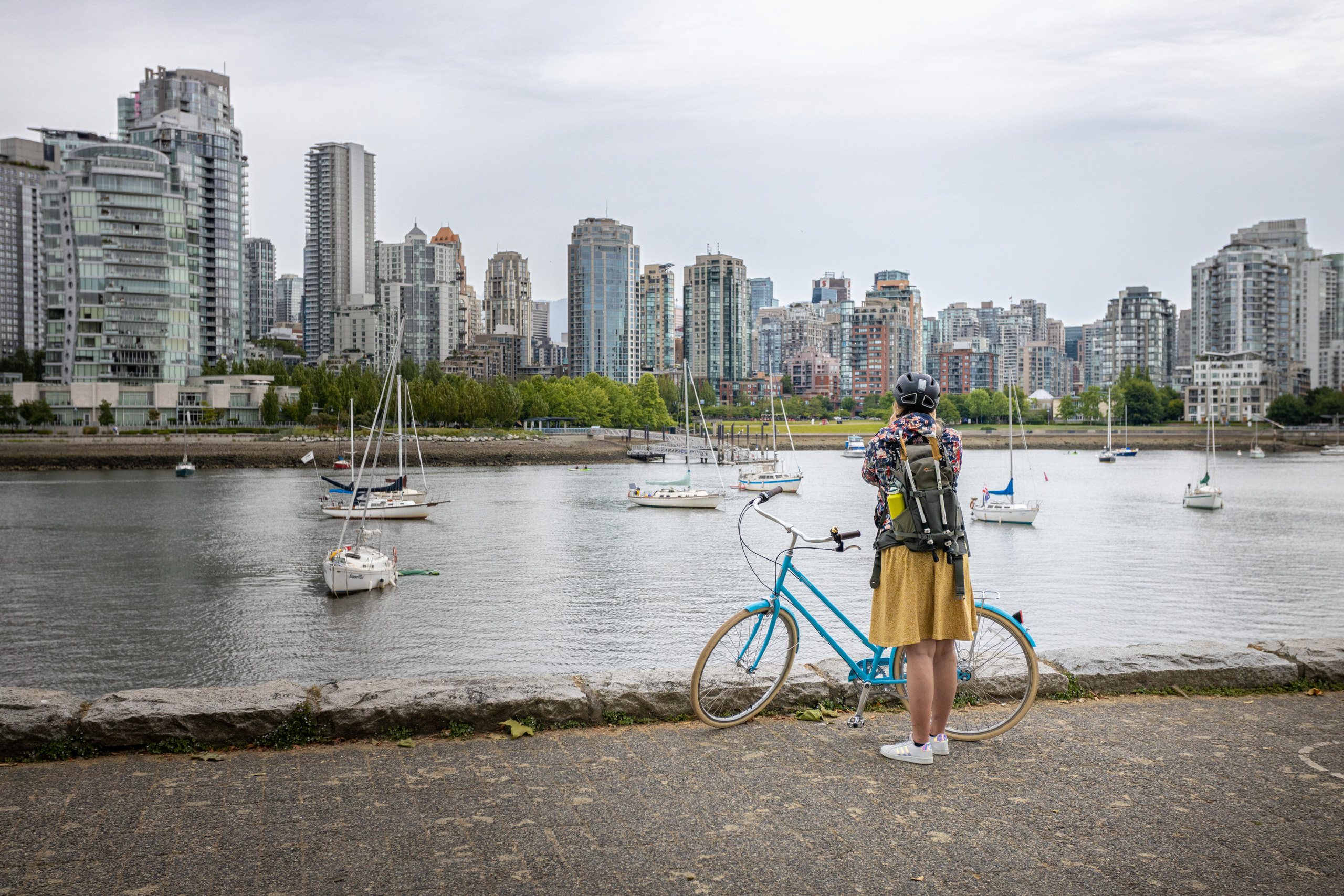 Manouk in een gele jurk en bloemenjas voor een blauwe fiets terwijl ze langs de waterkant een foto maakt van de skyline van Vancouver tijdens een camperreis door Canada