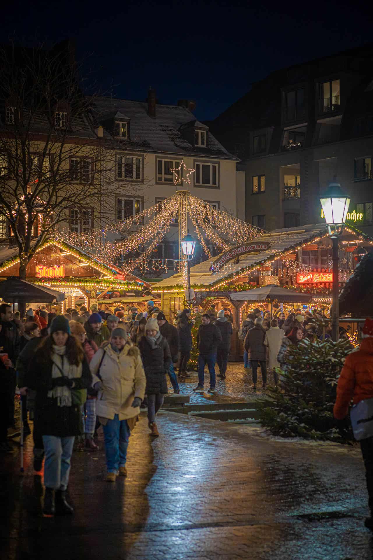 Kerstmarkt in Koblenz in het donker met mensen die rondlopen voor verlichte kramen. In het midden staat een paal waarvandaan snoeren lichtjes alle kanten op gaan, als een soort piramide.