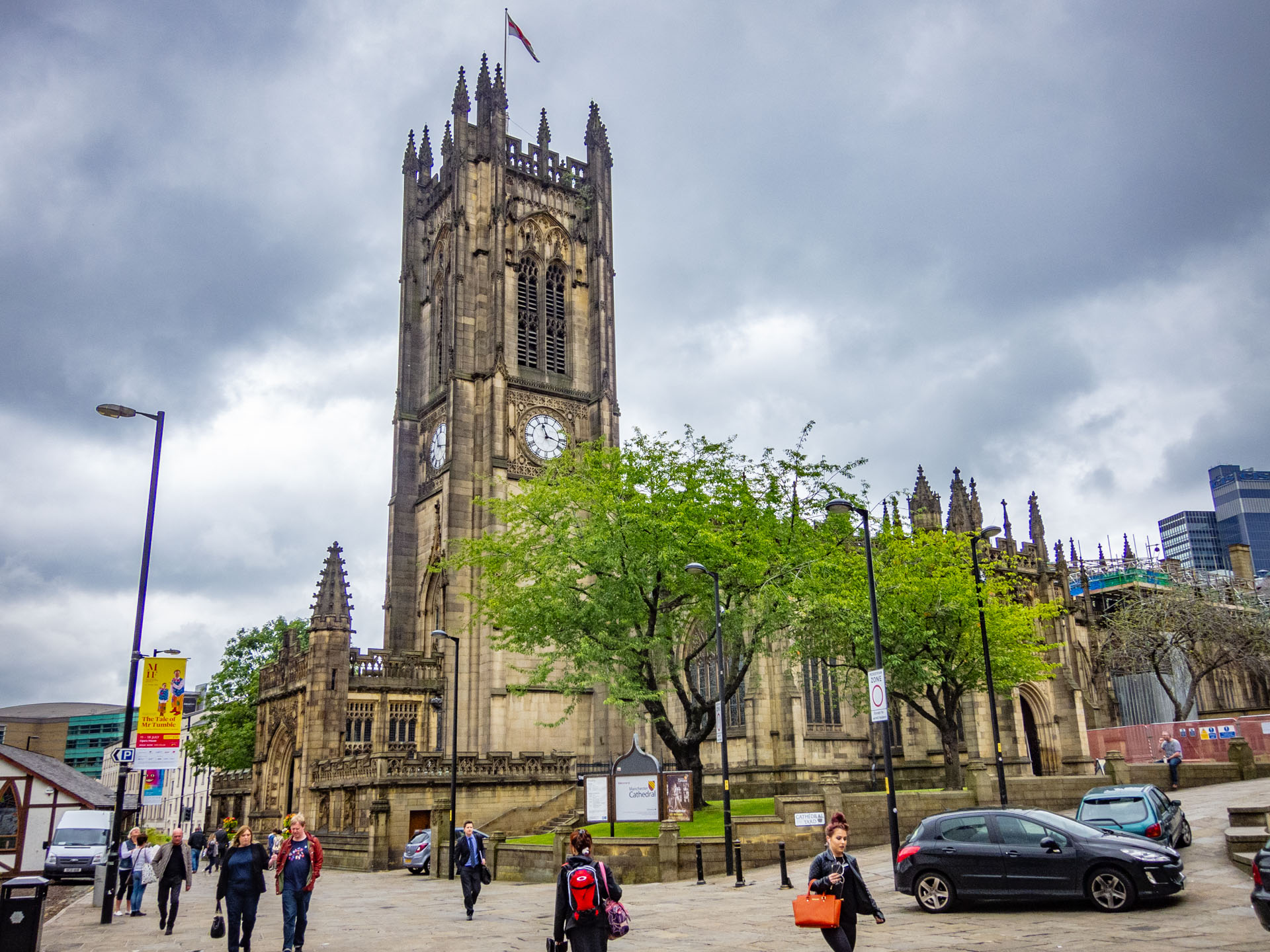 De kathedraal van Manchester met boom ervoor op een bewolkte dag, gezien vanaf overzijde van de straat