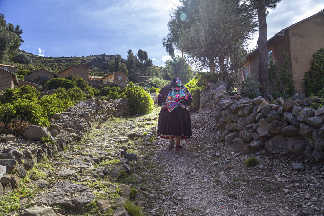 Vrouw in Peruaanse kledendracht loopt met rug naar de camera een heuvel op met een stenen muurtje rechts op het eiland Amantani in het Titicacameer in Peru