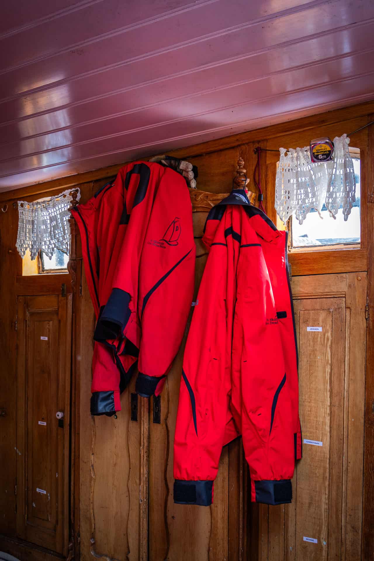 Rode jassen met logo van het Drachtster skûtsje hangen aan een haakje voor houten deuren in een skûtsje met vitrage voor de ramen