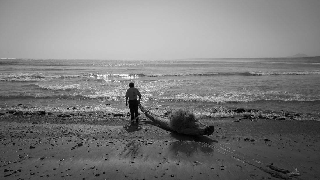 Zwart-witfoto van een man die een lange, rieten boot het water in trekt op het strand van Huanchaco in Peru