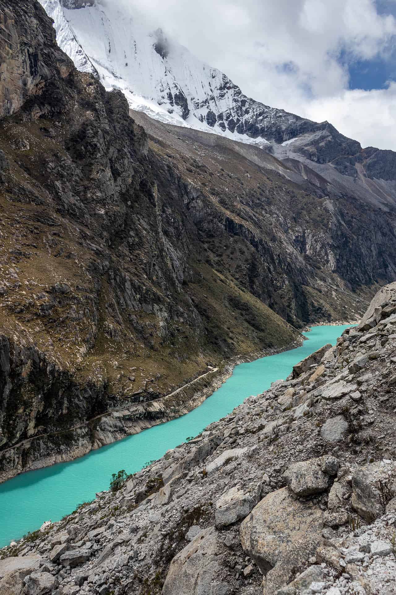 Felblauwe langwerpig meer Laguna Paron in Peru met rotsberg aan de rechterkant en hoge, besneeuwde bergen aan de linkerkant.