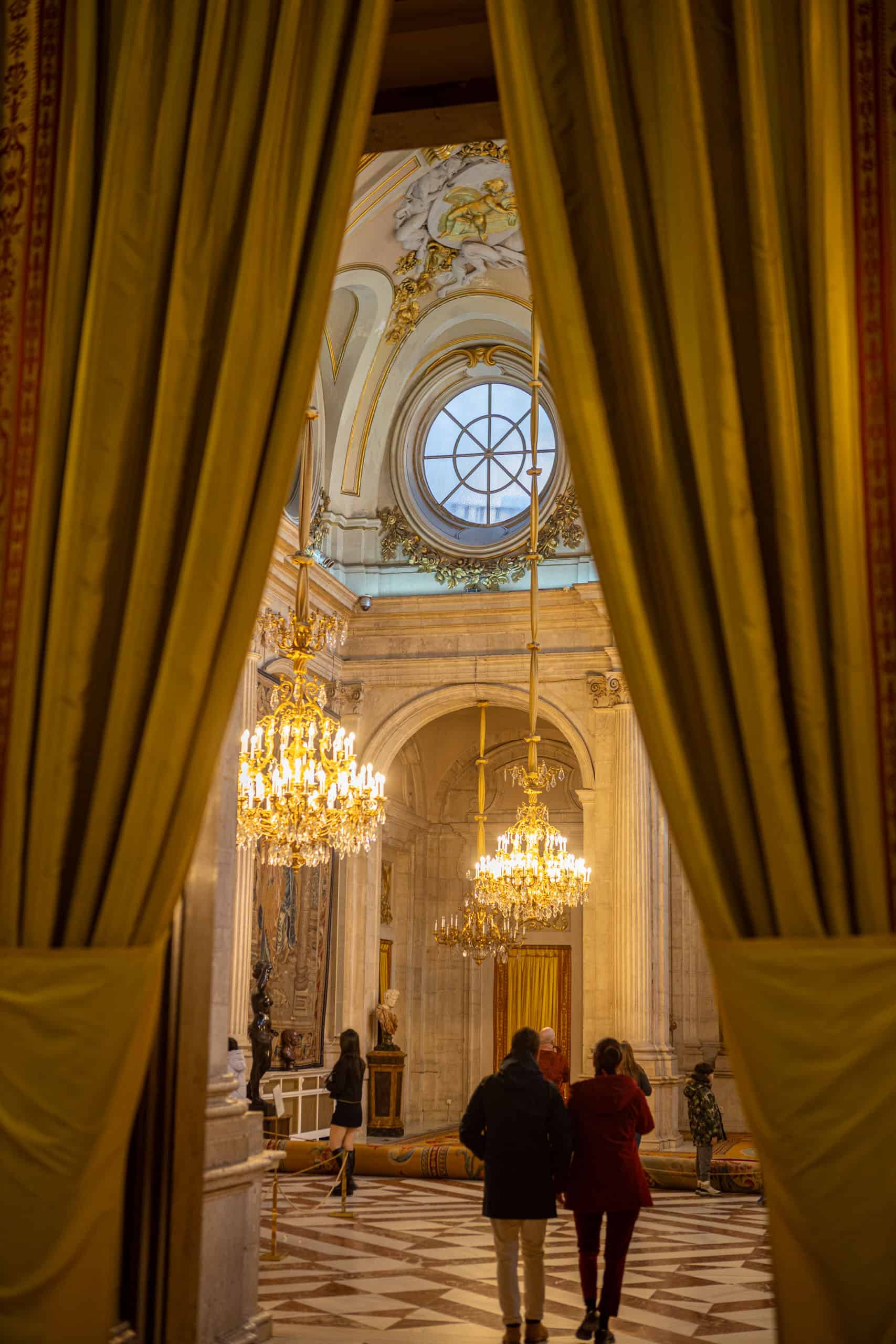 Ontvangstzaal in Palacio Real, gezien tussen de gordijnen van de zaal ervoor met rond raam en uitbundige kroonluchters