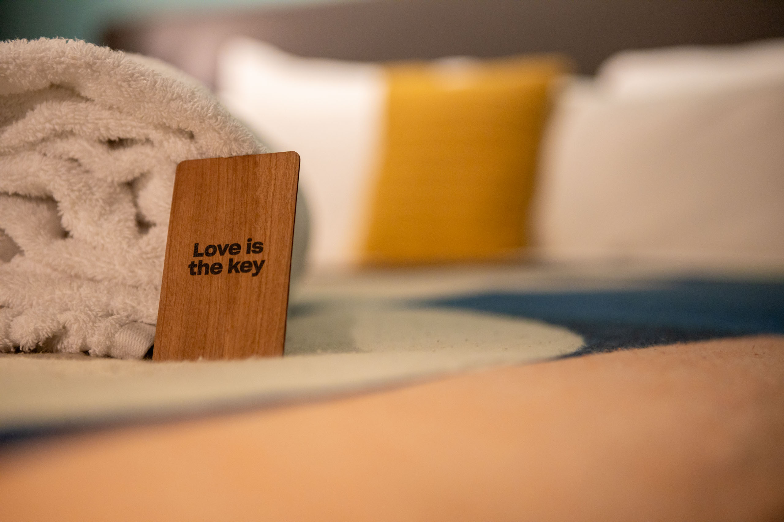 Bed met roze dekbed met erop opgerolde handdoeken en de kamersleutel, een houten pasje met 'Love is the key' erop.