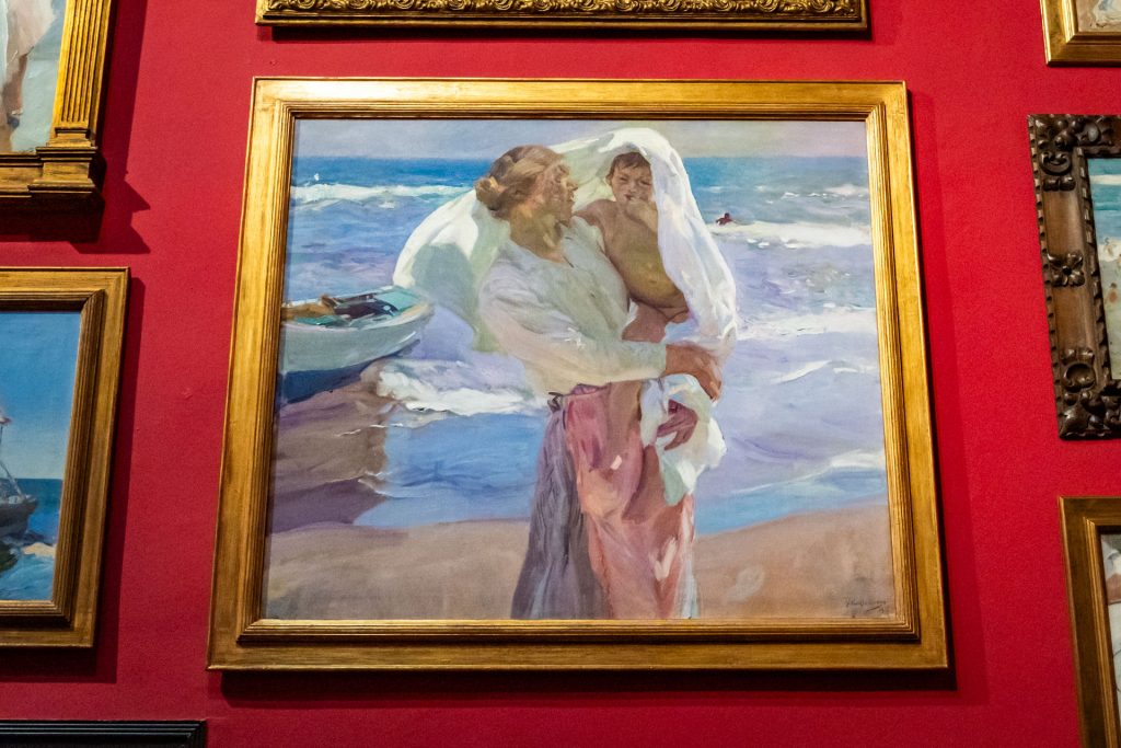 Schilderij Saliendo del baño of After Bathing van Sorolla hangt aan een rode muur in de studio in zijn woonhuis. Erop staat een vrouw met witte blouse met blauw/roze rok die een bloot kind vasthoudt waar een witte doek overheen ligt.
