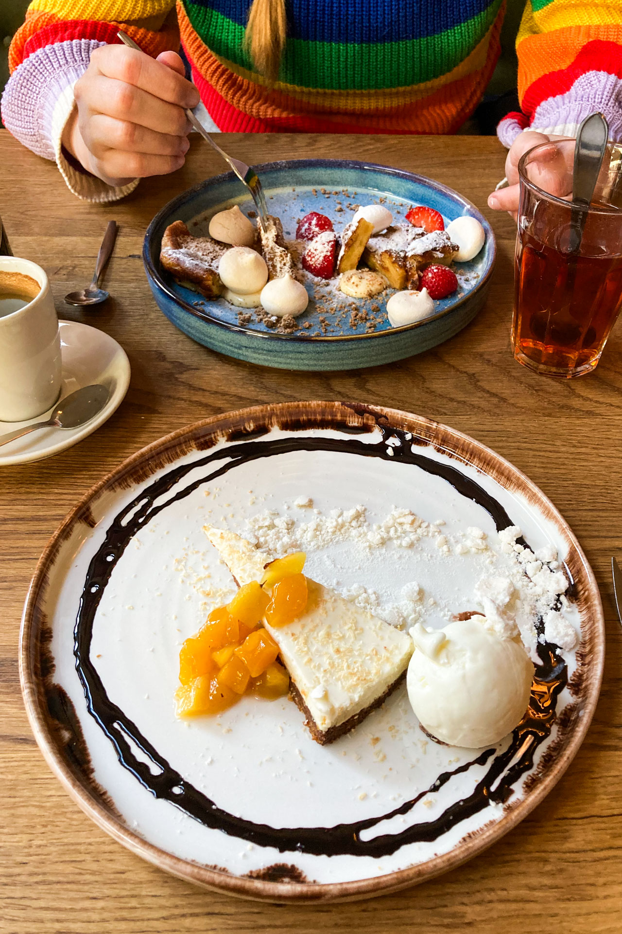 Vooraan een bord met cheesecake met ijs en geel fruit en achterop de tafel een bord met een dessert met allerlei onderdelen, waaronder merengue puntjes en aardbeien.