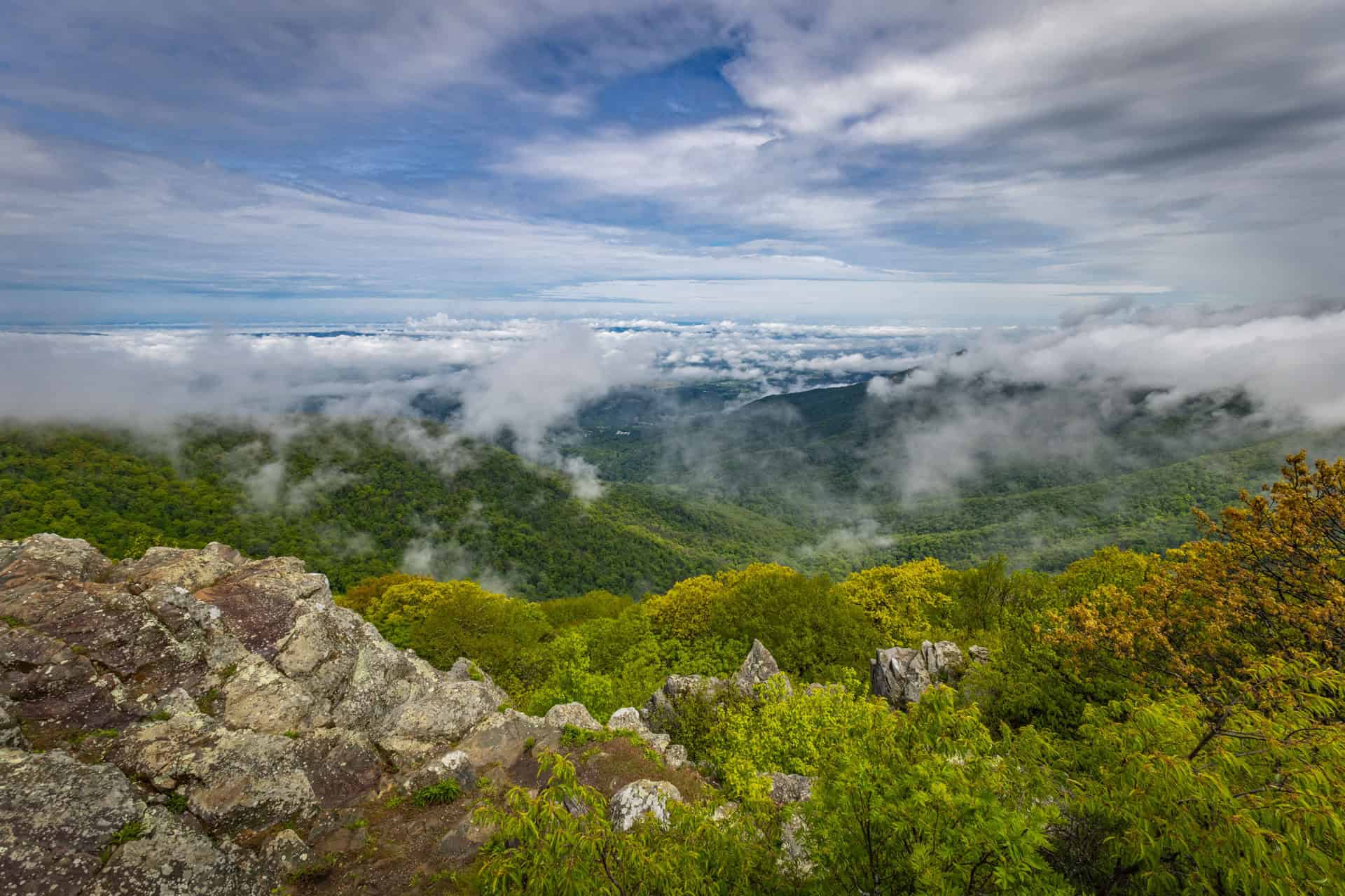 Uitzicht vanaf Hawksbill Mountain op golvende groene heuvels bedekt met mistflarden en wolken onder een deels bewolkte lucht. Op de voorgrond ruige rotspartijen en frisgroene bomen die het berglandschap omlijsten.