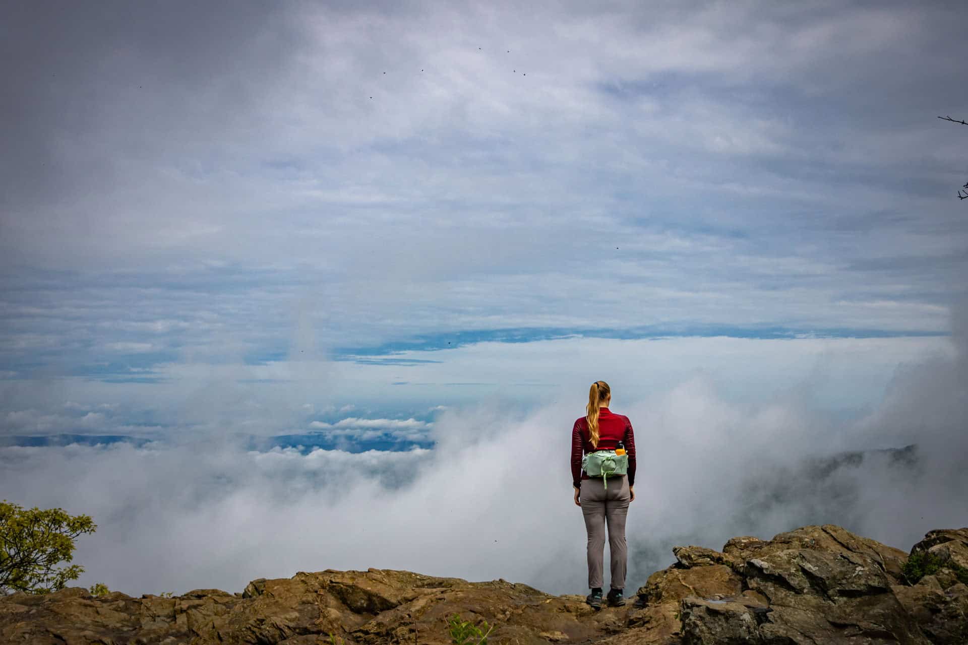 Vrouw met een lichtgroene heuptas en rode trui staat op de top van Hawksbill Mountain, uitkijkend over een zee van wolken en een dramatische, bewolkte lucht. De rotsachtige voorgrond benadrukt het ruige berglandschap.