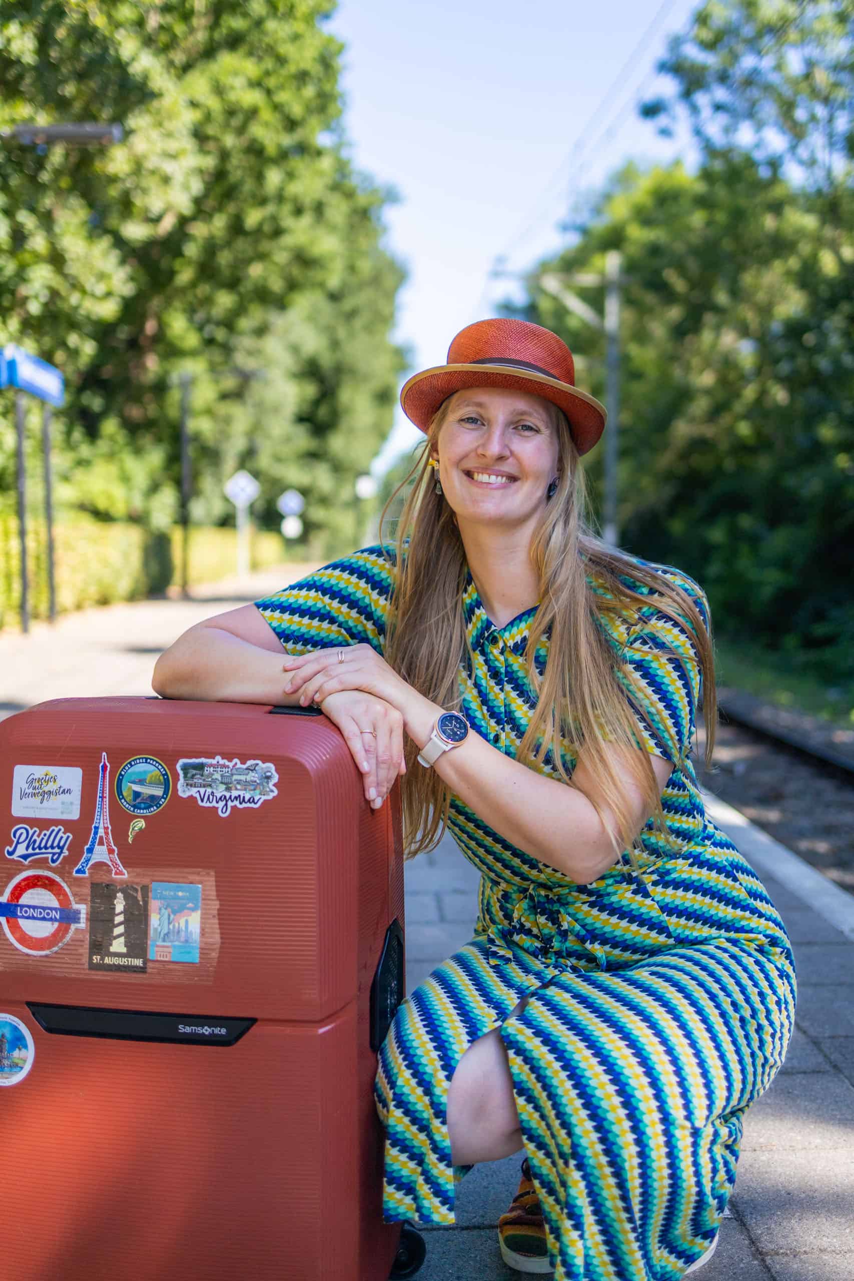 Vrolijke vrouw met lange blonde haren en oranje hoed leunt op een rode koffer vol reisstickers, wachtend op een perron. Perfecte sfeerbeeld bij een paklijst vliegvakantie, met zomerse kleding en glimlach die reisplezier uitstraalt.